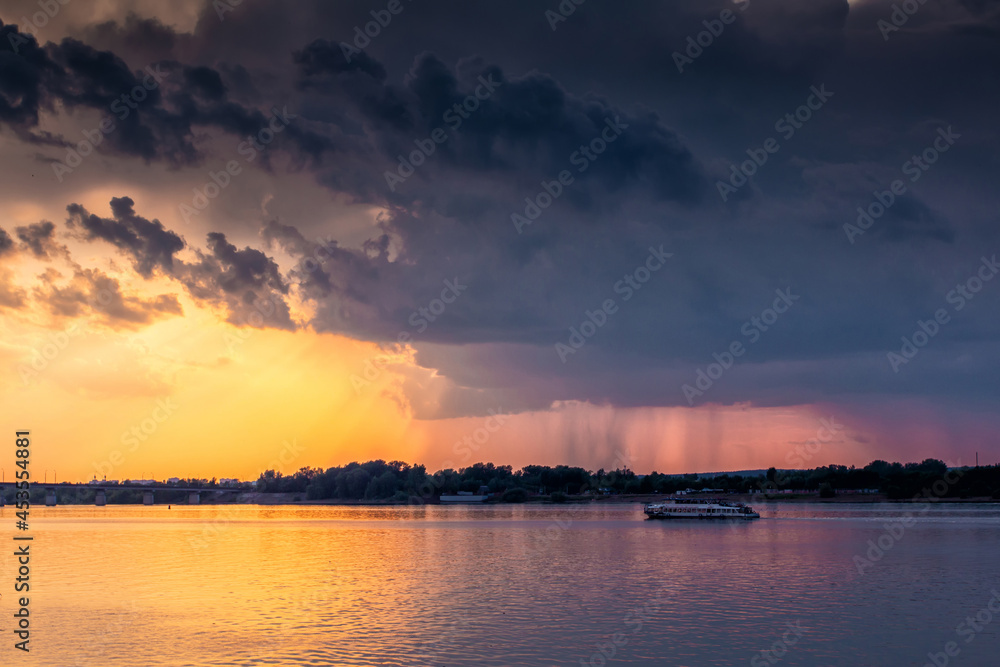 A small ship is sailing along the river against the backdrop of the sunset. Rain clouds in the sky. It's raining in the distance