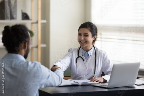 Happy young female indian ethnicity general practitioner physician therapist shaking hands with african american male patient, getting acquainted or making agreement at meeting in clinic office.