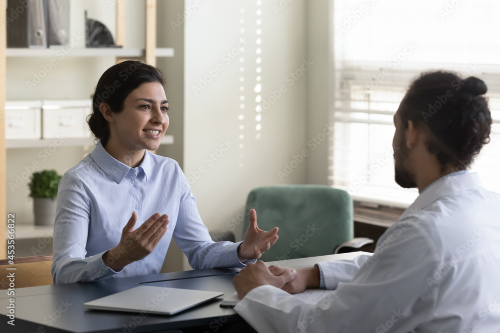 Obraz premium Smiling beautiful young indian ethnicity patient talking with professional african biracial family doctor therapist physician, discussing illness treatment or vaccination at checkup meeting in clinic.