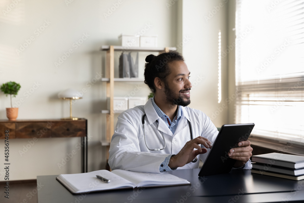 Dreamy young african american gp doctor physician using digital computer tablet, looing in distance distracted from remote work, web surfing useful information or consulting patient remotely. Dreamy young african american gp doctor physician using digital computer tablet, looing in distance distracted from remote work, web surfing useful information or consulting patient remotely.