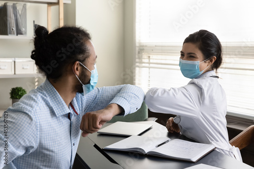 Happy young mixed race male patient and female gp doctor in facial protective masks touching elbows, starting or finishing checkup meeting in clinic, avoiding hand contact, covid prevention cocept.