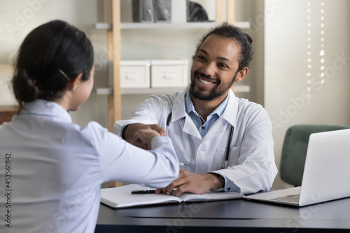 Smiling handsome millennial african american gp doctor physician shaking hands with indian female patient, getting acquainted or making agreement at regular checkup meeting in clinic office room. © fizkes