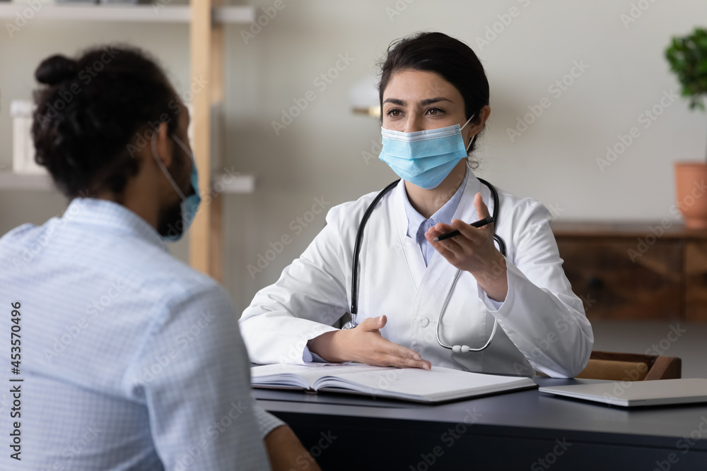 Φωτογραφία Concentrated young indian ethnic female gp doctor in facial protective mask giving professional healthcare consultation or explaining disease treatment to african american patient in clinic office