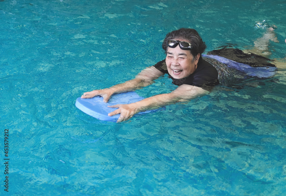 happy and healthy Asian senior woman swimming with kickboard in a swimming pool. Smiling old woman swimming with inflatable board in swimming pool.  Elderly active lifestyle.