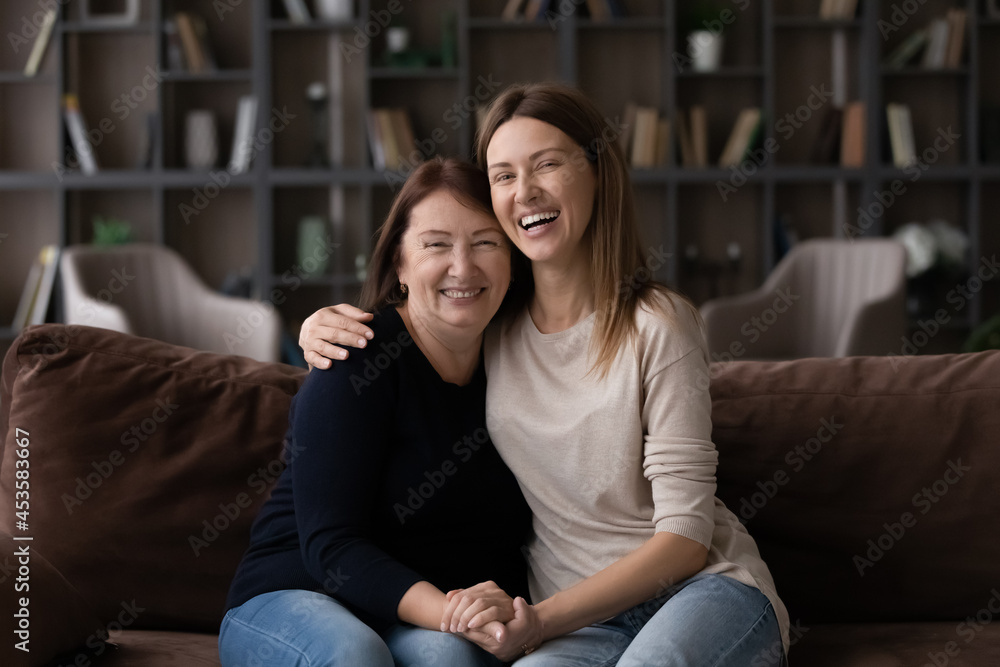 Portrait of smiling affectionate young woman cuddling loving middle aged mature retired mother, resting together on comfortable couch. Joyful multigenerational female family enjoying relaxed pastime. Portrait of smiling affectionate young woman cuddling loving middle aged mature retired mother, resting together on comfortable couch. Joyful multigenerational female family enjoying relaxed pastime.