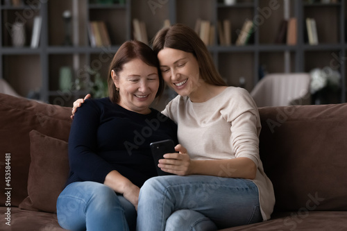 Smiling affectionate young woman showing photos or video on smartphone to loving middle aged elderly retired mother, having fun using modern technology gadget relaxing together on comfortable sofa.
