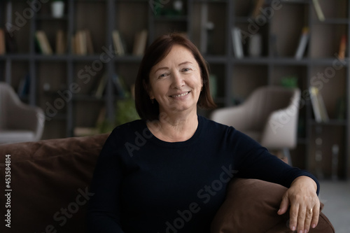 Portrait of happy confident sincere elderly senior woman resting on comfortable sofa in stylish living room. Joyful smiling candid middle aged retires grandmother relaxing on cozy couch at home.