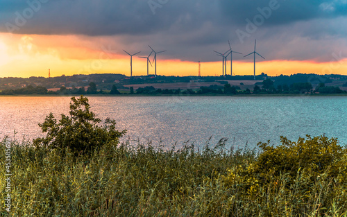 Wind farm on a hill above the lake at sunrise, thickets on the shores of the lake in the foreground; Darłowo, Poland, Baltic Sea
