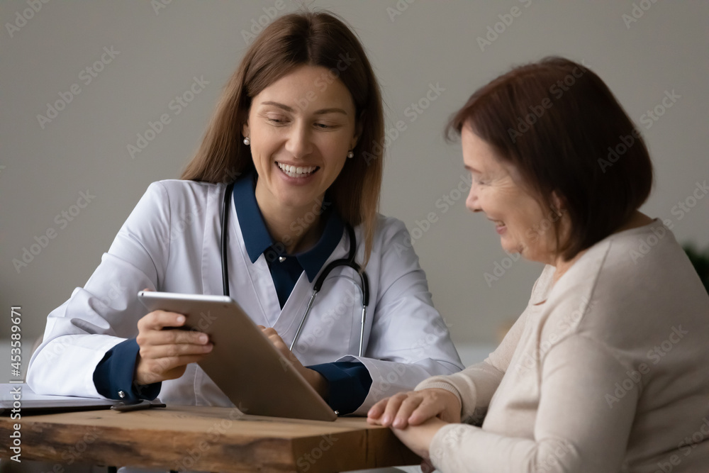 Happy young female GP doctor physician showing health test results on digital computer tablet to happy old mature retired woman, feeling satisfied with disease treatment, medicine and modern tech. Happy young female GP doctor physician showing health test results on digital computer tablet to happy old mature retired woman, feeling satisfied with disease treatment, medicine and modern tech.