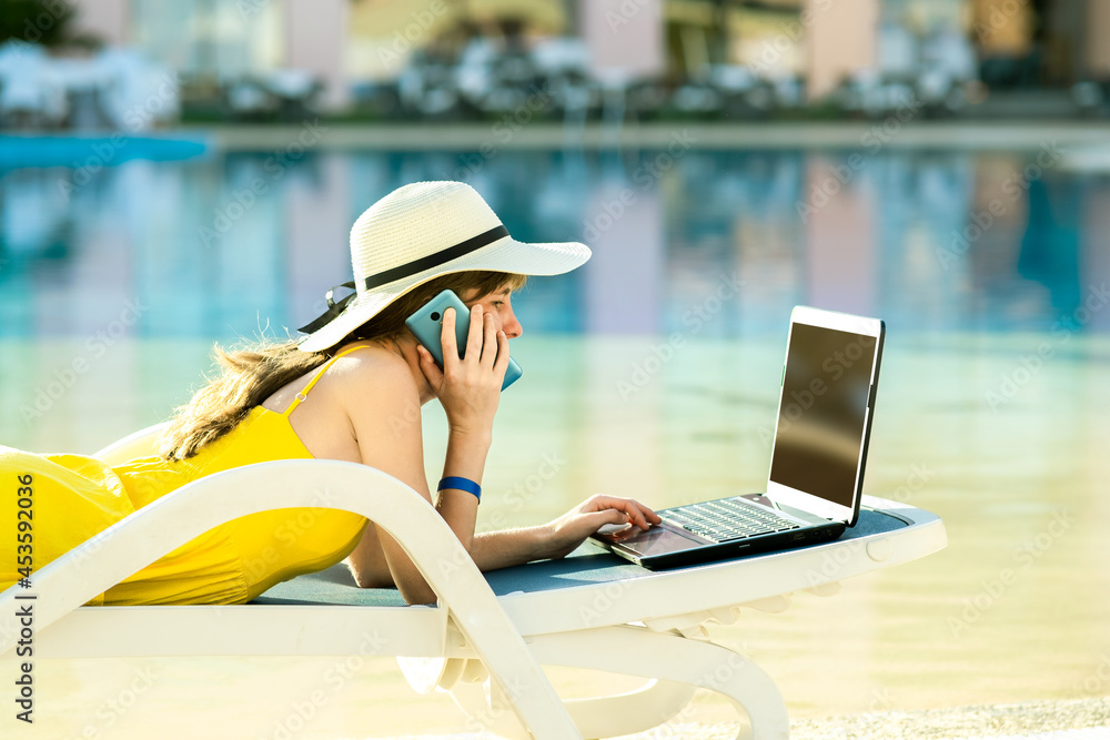 Young woman on beach chair at swimming pool working on computer laptop and talking on sell phone in summer resort. Remote work and freelance job while travelling concept.