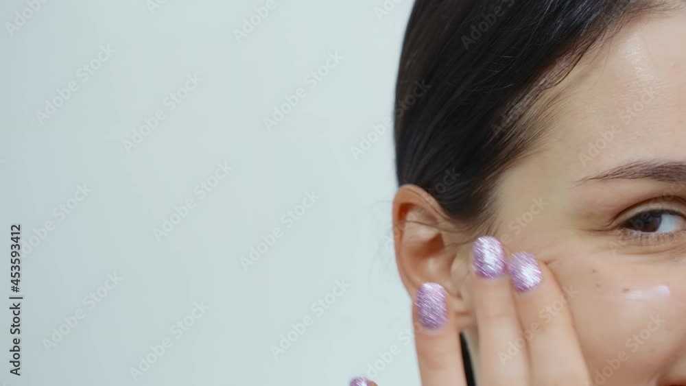 Close up beautiful woman with brown hair and purple manicure applying cream on face on white background. Concept of process of skin care on face in bathroom. Close up beautiful woman with brown hair and purple manicure applying cream on face on white background. Concept of process of skin care on face in bathroom.