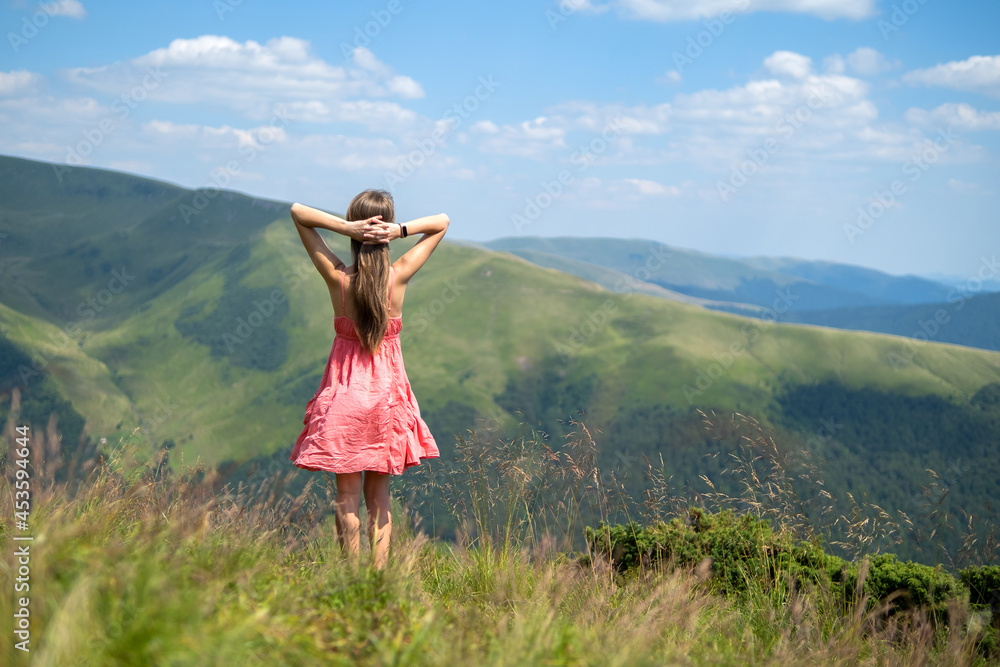 Rear view of a happy woman hiker in red dress standing on grassy hill on a windy day in summer mountains with outstretched arms enjoying view of nature.