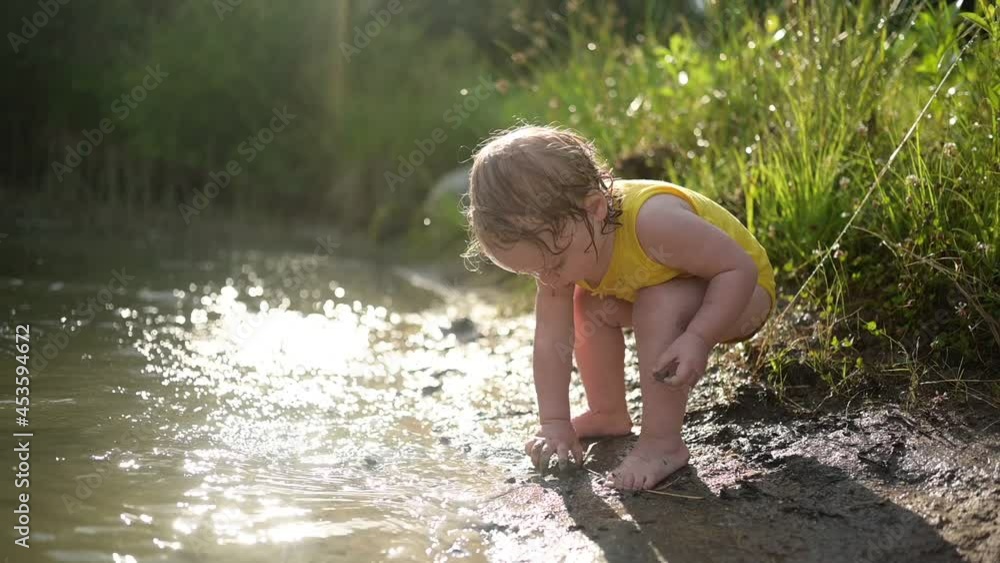 Little funny cute blonde girl child toddler in yellow wet bodysuit playing by the lake waterside shore at sunset outside. Baby with mud on hands and legs. Water activities at summer Little funny cute blonde girl child toddler in yellow wet bodysuit playing by the lake waterside shore at sunset outside. Baby with mud on hands and legs. Water activities at summer