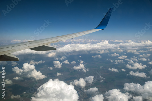 View of jet airplane wing from inside flying over white puffy clouds in blue sky. Travel and air transportation concept.