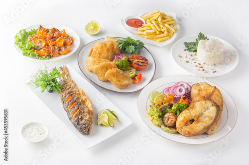 Assorted fried fish dishes - whole trout and large pieces with baked vegetables, rice, herbs and lemon on a white table