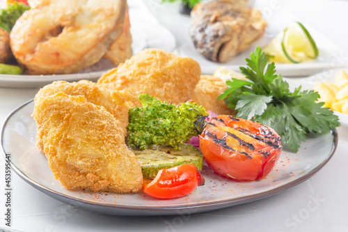 Large chunks of fried fish - pike perch, breaded in breadcrumbs with herbs, red onion, lemon and baked vegetables in a white plate. Close-up
