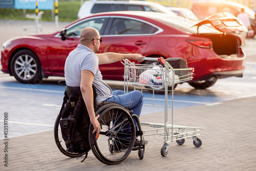 Adult disabled man in a wheelchair pushes a cart towards a car in a supermarket parking lot © romaset