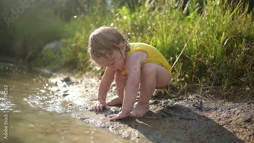 Little funny cute blonde girl child toddler in yellow wet bodysuit playing by the lake waterside shore at sunset outside. Baby with mud on hands and legs. Water activities at summer