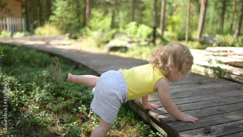 Little funny cute blonde girl child toddler in yellow undershirt and grey shorts walking playing on wooden eco trail outside at summer at front yard countryside. Healthy happy childhood concept. Little funny cute blonde girl child toddler in yellow undershirt and grey shorts walking playing on wooden eco trail outside at summer at front yard countryside. Healthy happy childhood concept.
