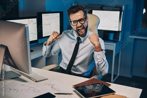 Happy young man gesturing and looking at cmputer while staying late in the office photo