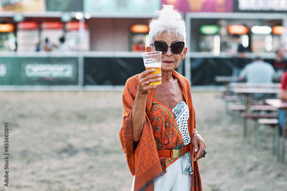 A grandma standing outdoor at a music festival and showing her beer. She is having a good time. - obrazy, fototapety, plakaty 