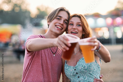 A young couple is standing outdoor at a music festival and toasting with a fresh cold beer. Summer is calling.