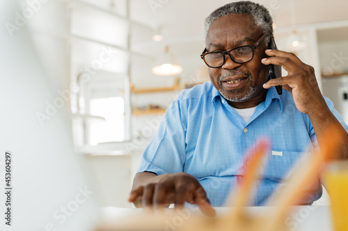 An old black man is sitting at his home office, talking on the phone with a customer support worker and using his laptop.