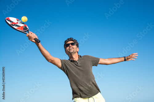 Man Holding A Paddle Tennis Racket Hitting The Ball On A Blue Background. Young Sportsman Playing Tennis On The Beach. Paddle Player © Nanci