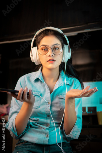 Low angle image of bespectacled Asian, Indian young woman in denim shirt sitting at desktop and looking at the camera with a shock expression while listening music through headphones at home. 
