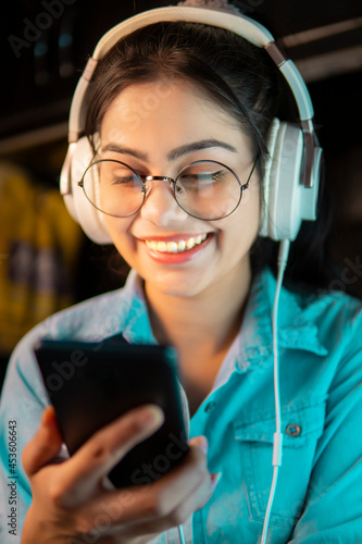 Low angle image of Beautiful happy bespectacled Asian, Indian young woman in denim shirt hearing music through headphones and using a smartphone.