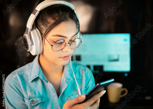 Beautiful happy bespectacled Asian, Indian young woman in denim shirt sitting at desktop and listening music through headphones and using a smartphone while taking break from work at home. 