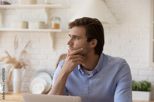 Pensive young caucasian business man looking in distance considering problem solution, thinking on new creative project, working on computer alone in modern kitchen, distant freelance workday concept.