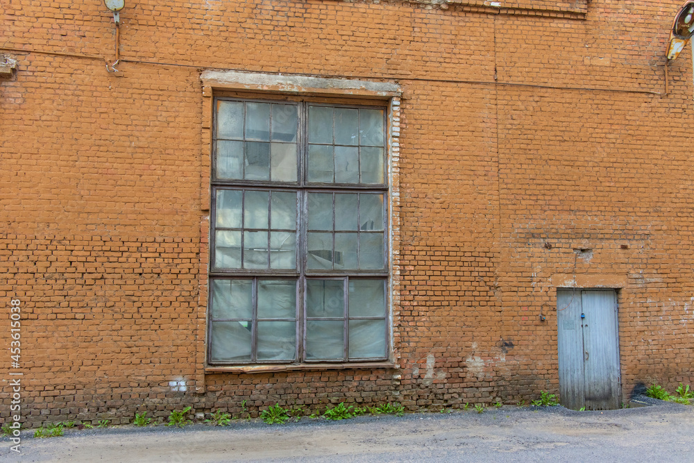 A large old dilapidated red brick house with a large dilapidated window and a gray wooden door. An old abandoned factory building.