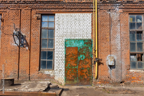 A large old dilapidated house of red and white bricks with large dilapidated windows and a multi-colored metal door. Old dirty abandoned factory building.