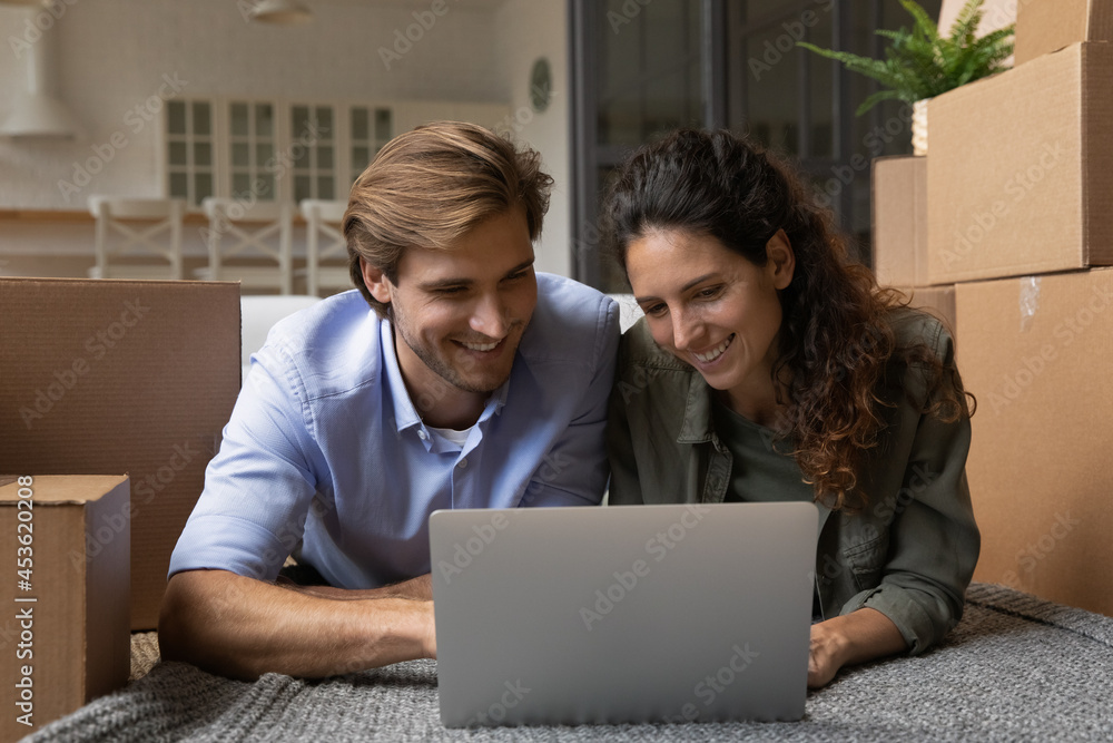 Happy young family couple lying on floor between cardboard boxes, web surfing information online on computer, shopping choosing goods in internet store, planning purchasing interior decorations.