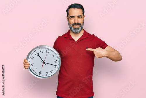 Middle aged man with beard holding big clock pointing finger to one self smiling happy and proud