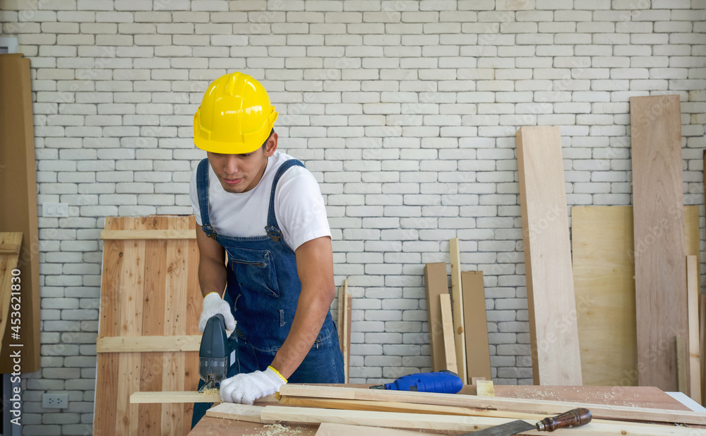 Asian carpenter wearing a yellow hardhat, use electric hand saw to cut plywood at the woodworking facility. A desk full of hand tools and wood piles. Morning work atmosphere in the workshop room.