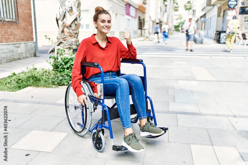 Young brunette woman sitting on wheelchair outdoors pointing thumb up to the side smiling happy with open mouth Young brunette woman sitting on wheelchair outdoors pointing thumb up to the side smiling happy with open mouth