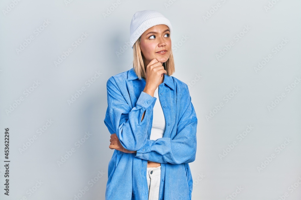 Beautiful blonde woman wearing wool hat with hand on chin thinking about question, pensive expression. smiling and thoughtful face. doubt concept. Beautiful blonde woman wearing wool hat with hand on chin thinking about question, pensive expression. smiling and thoughtful face. doubt concept.