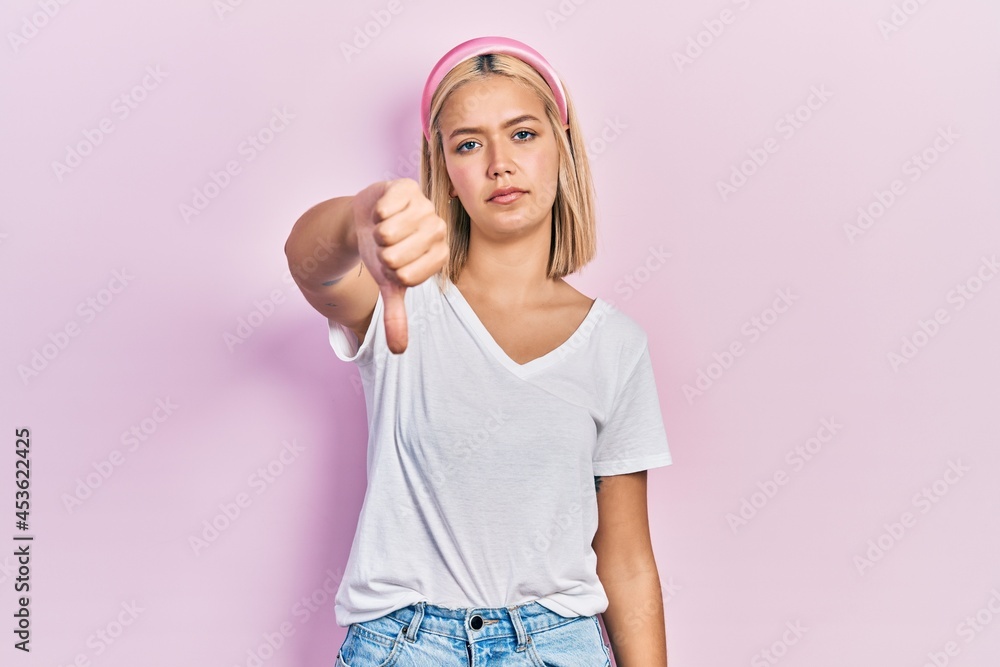 Beautiful blonde woman wearing casual white t shirt looking unhappy and angry showing rejection and negative with thumbs down gesture. bad expression. Beautiful blonde woman wearing casual white t shirt looking unhappy and angry showing rejection and negative with thumbs down gesture. bad expression.