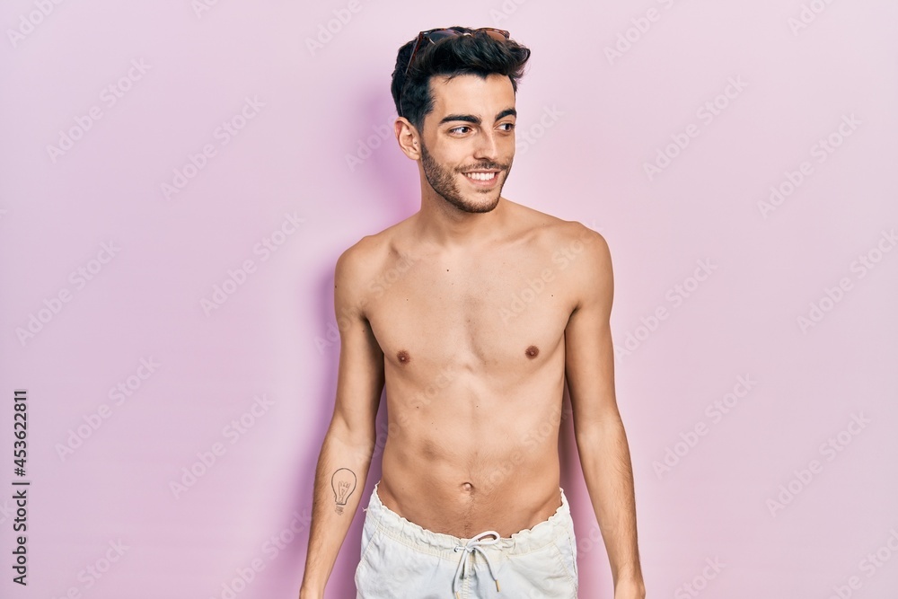 Young hispanic man wearing swimwear shirtless looking away to side with smile on face, natural expression. laughing confident. Young hispanic man wearing swimwear shirtless looking away to side with smile on face, natural expression. laughing confident.