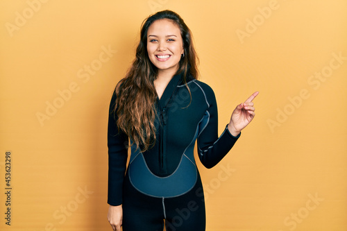 Young hispanic girl wearing diver neoprene uniform with a big smile on face, pointing with hand finger to the side looking at the camera. photo