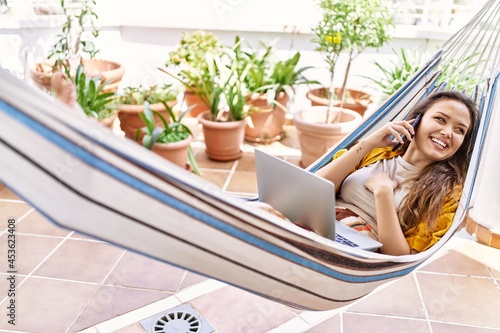 Young hispanic girl talking on the smartphone and using laptop lying on hammock at the terrace.