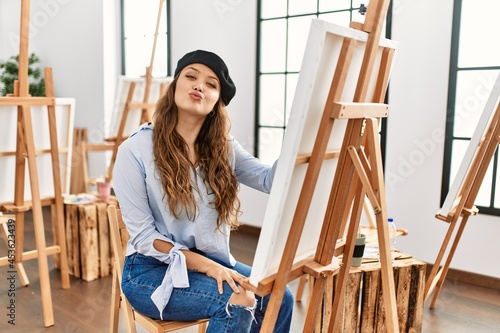 Young hispanic artist woman painting on canvas at art studio looking at the camera blowing a kiss on air being lovely and sexy. love expression.