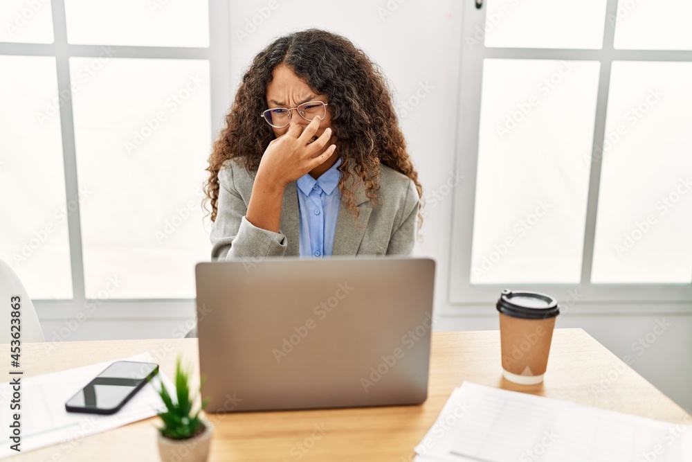 Beautiful hispanic business woman sitting on desk at office working with laptop smelling something stinky and disgusting, intolerable smell, holding breath with fingers on nose. bad smell Beautiful hispanic business woman sitting on desk at office working with laptop smelling something stinky and disgusting, intolerable smell, holding breath with fingers on nose. bad smell