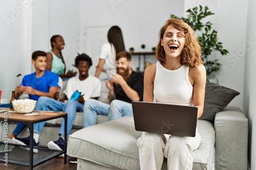 Group of young friends smiling happy sitting on the sofa. Woman using laptop at home.