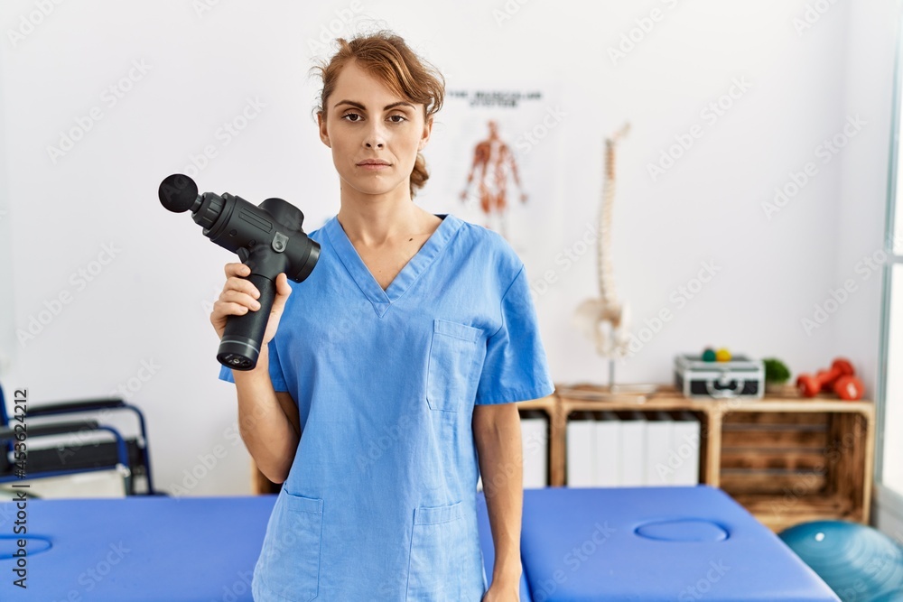 Beautiful caucasian woman holding therapy massage gun at wellness center thinking attitude and sober expression looking self confident