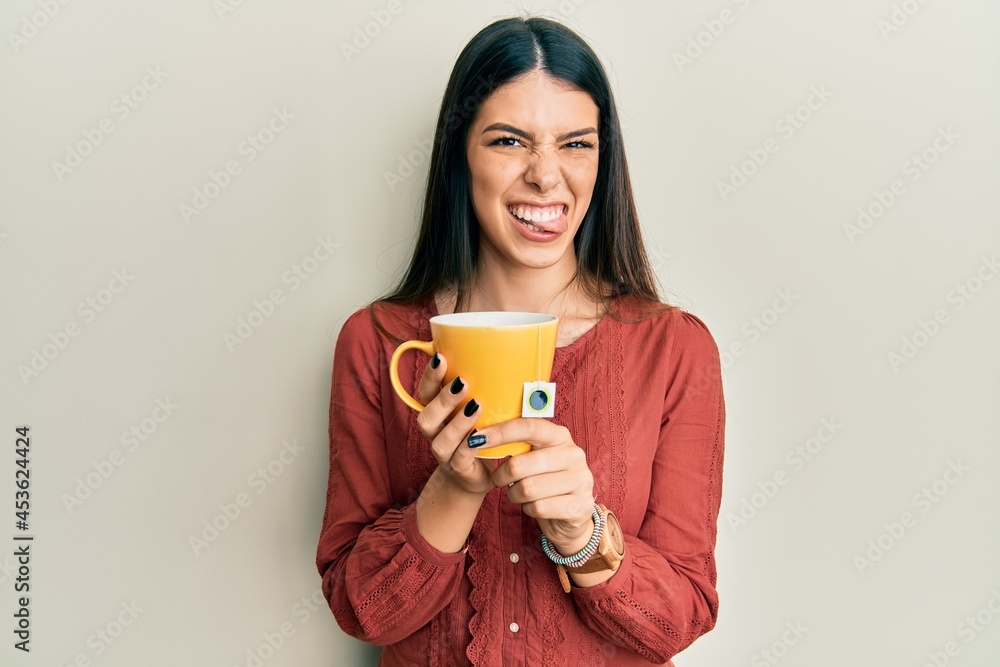 Young hispanic woman holding cup of tea sticking tongue out happy with funny expression. Young hispanic woman holding cup of tea sticking tongue out happy with funny expression.