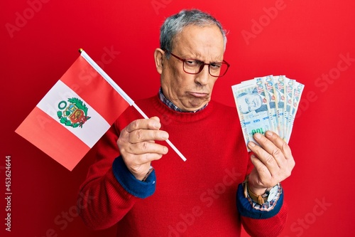 Handsome senior man with grey hair holding peru flag and peruvian sol banknotes depressed and worry for distress, crying angry and afraid. sad expression.