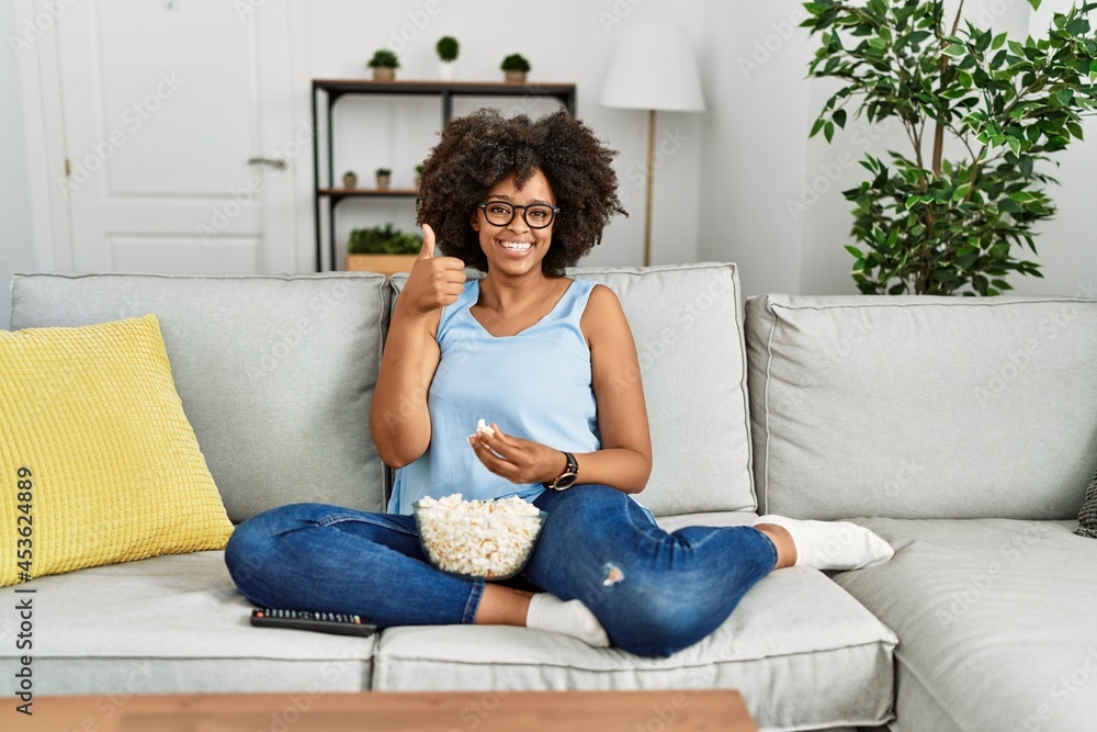 African american woman with afro hair sitting on the sofa eating popcorn at home smiling happy and positive, thumb up doing excellent and approval sign